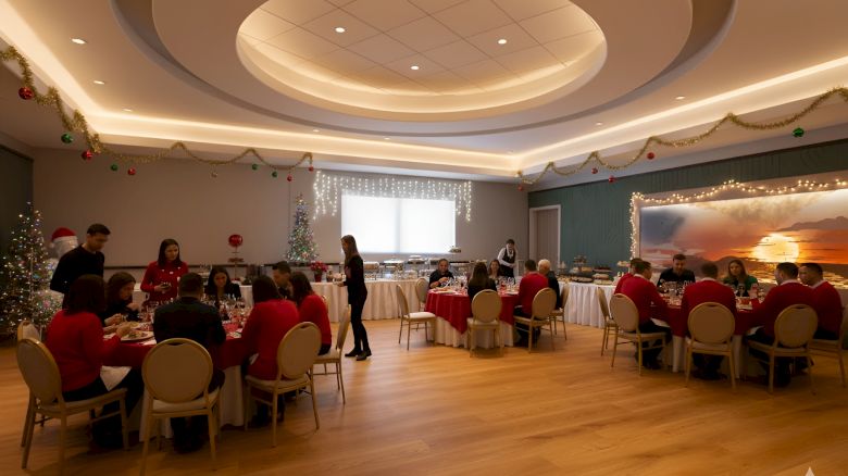 A festive gathering in a decorated room with tables, people dining, and Christmas decor like garlands and a Christmas tree.