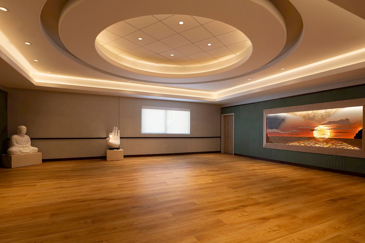 A serene room with a wooden floor, Buddha statue, large hand sculpture, and a wall display of a sunset over the ocean, under soft lighting.