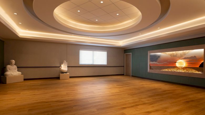 A serene room with a wooden floor, Buddha statue, large hand sculpture, and a wall display of a sunset over the ocean, under soft lighting.