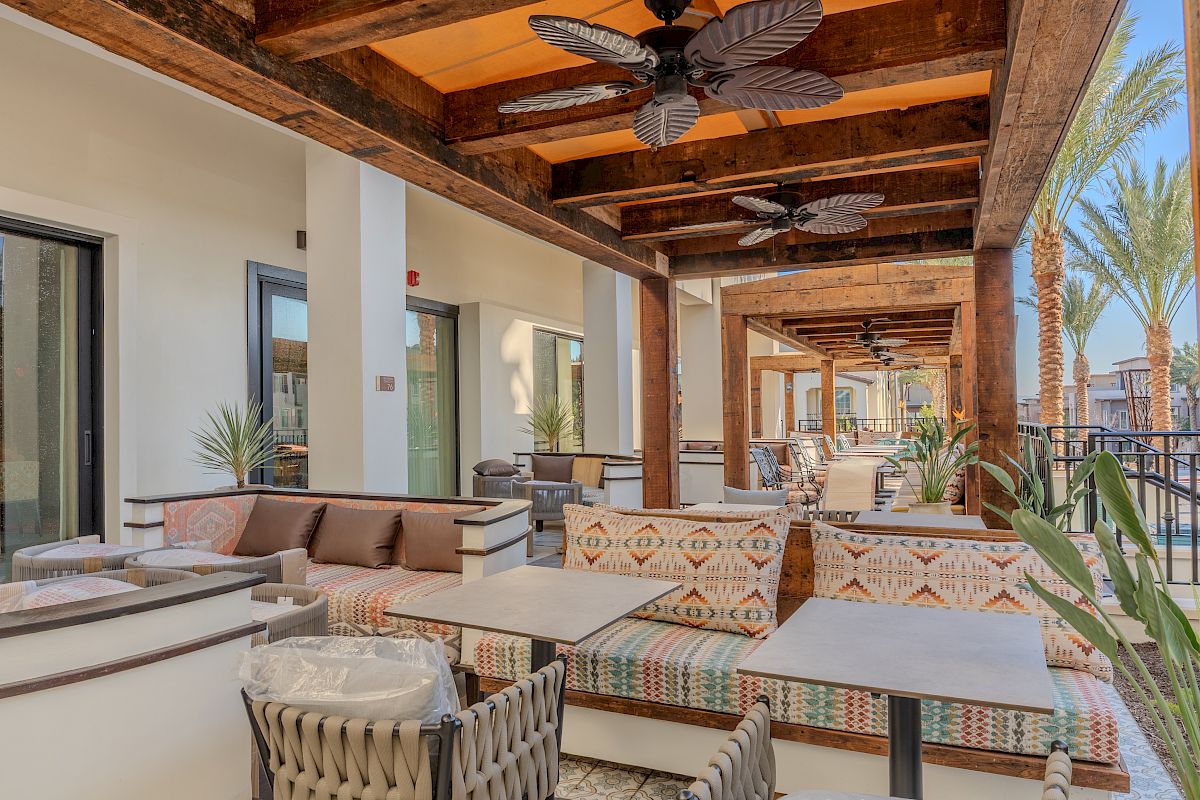 Outdoor seating area with beige sofas, patterned cushions, and tables under a wooden pergola. Ceiling fans and palm trees are visible.