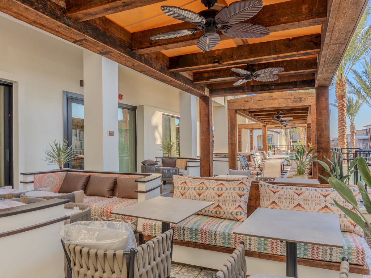 Outdoor seating area with beige sofas, patterned cushions, and tables under a wooden pergola. Ceiling fans and palm trees are visible.