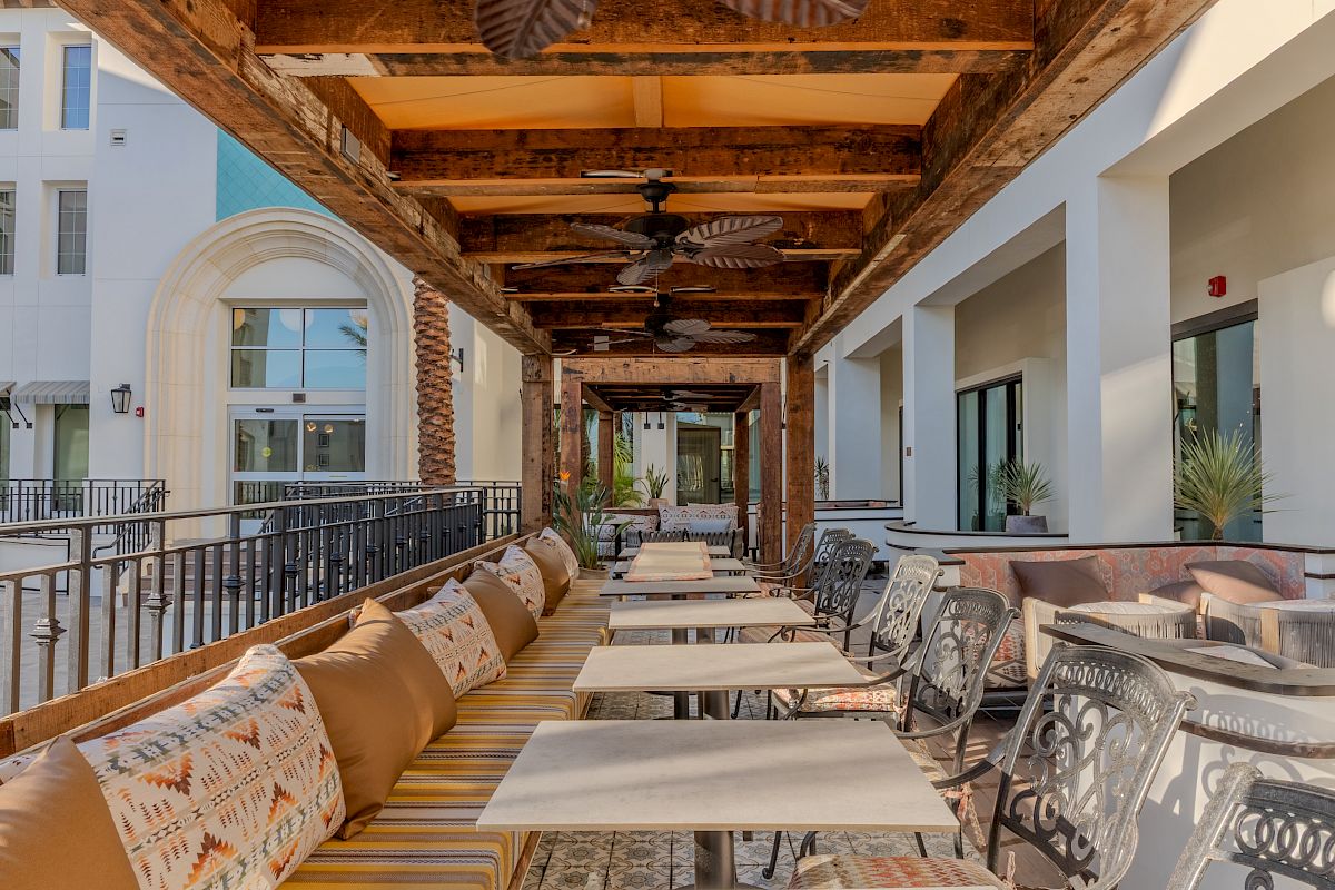 Outdoor terrace with tables, ornate chairs, cushioned bench seating, and wooden beams under a covered patio. Fans line the ceiling.