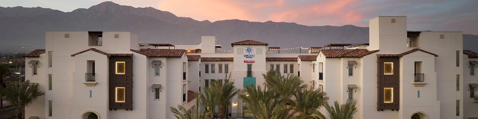 A large white building with palm trees in front, set against a mountain backdrop under a colorful sunset sky.