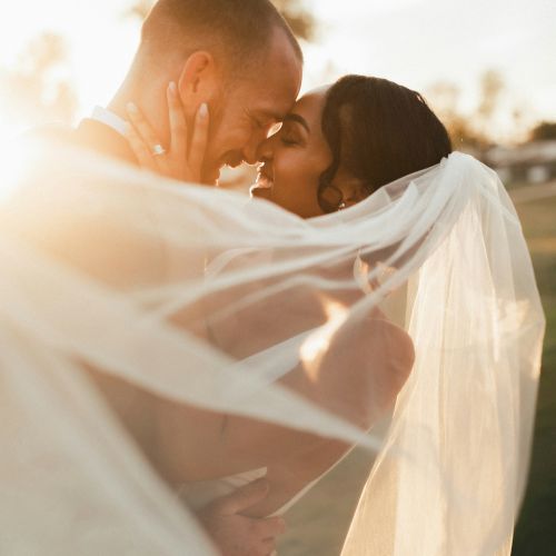 A bride and groom embrace, sharing a kiss outdoors with the sun setting behind them, the bride's veil flowing around them.