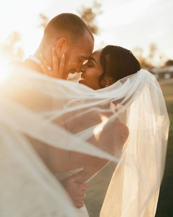 A bride and groom embrace, sharing a kiss outdoors with the sun setting behind them, the bride's veil flowing around them.