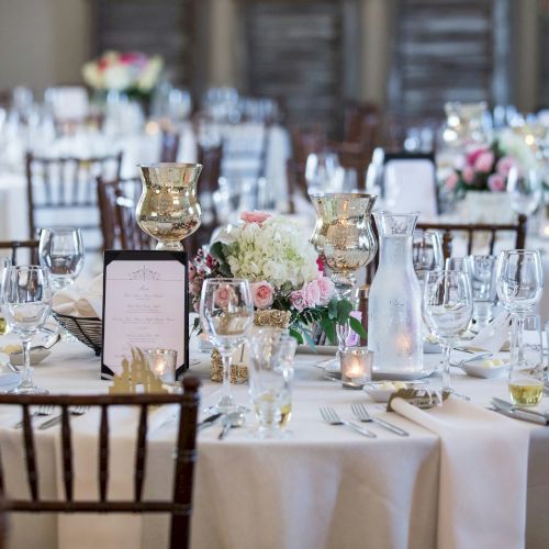 The image shows an elegantly set dining table with glassware, candles, flowers, and menu cards arranged for a formal event or wedding reception.