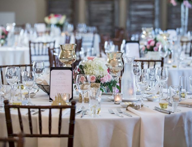 The image shows an elegantly set dining table with glassware, candles, flowers, and menu cards arranged for a formal event or wedding reception.