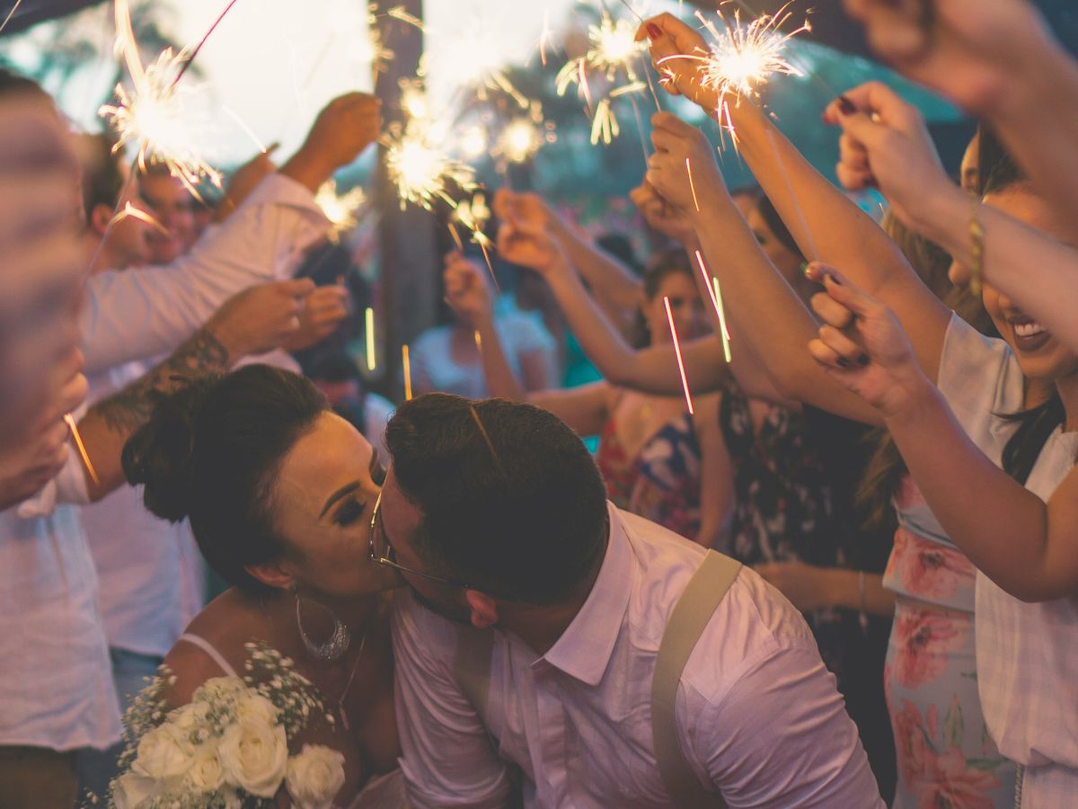 A couple is kissing amidst a crowd holding sparklers, suggesting a celebratory event, possibly a wedding or party, with a festive atmosphere.