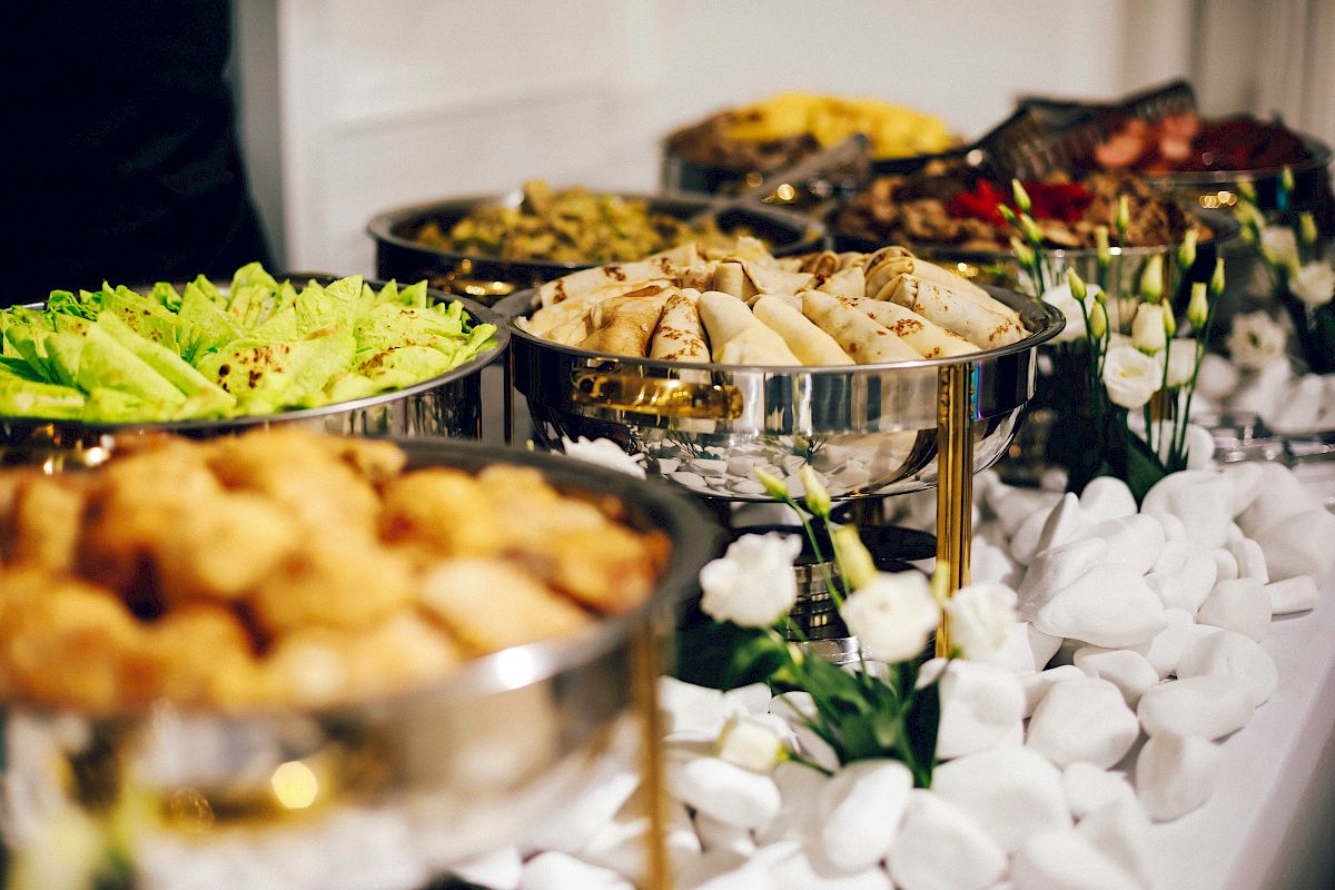 The image shows a buffet table with various dishes in metal trays, including salads, spring rolls, and fried items, arranged attractively.