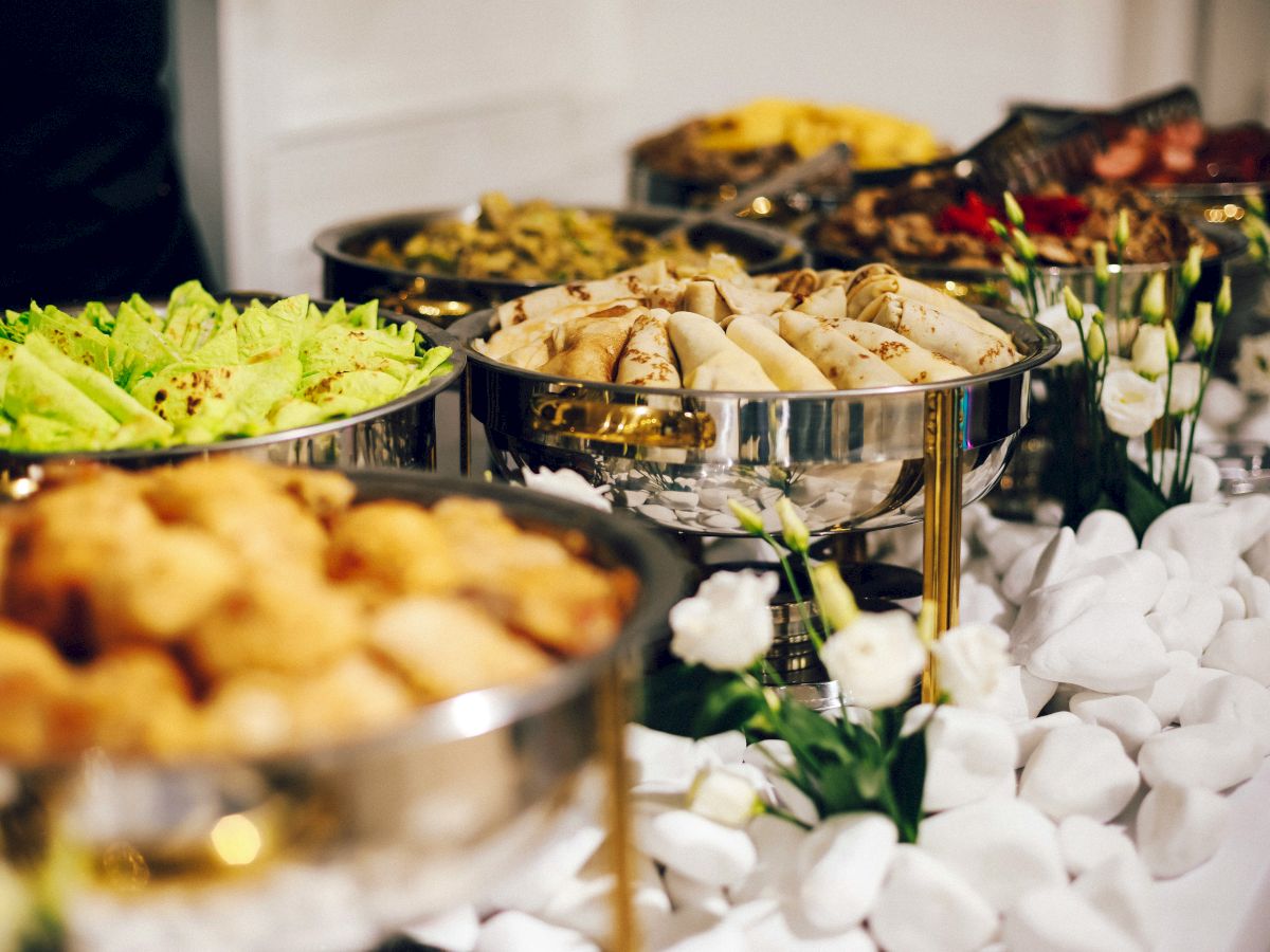 The image shows a buffet table with various dishes in metal trays, including salads, spring rolls, and fried items, arranged attractively.