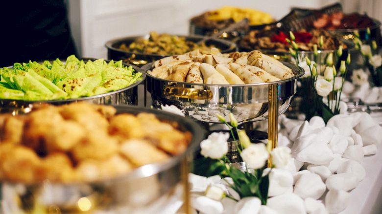 The image shows a buffet table with various dishes in metal trays, including salads, spring rolls, and fried items, arranged attractively.