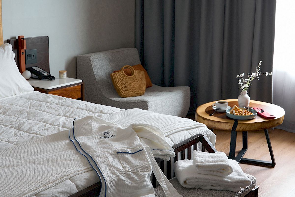 A cozy bedroom with a neatly made bed, robes, and towels. There's a chair, a small table with a vase, and a curtain by the window.