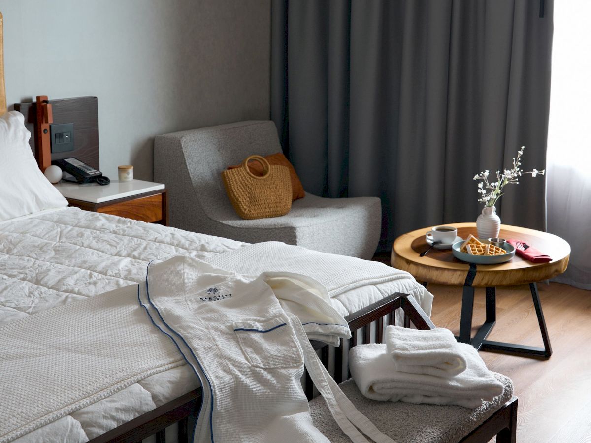 A cozy bedroom with a neatly made bed, robes, and towels. There's a chair, a small table with a vase, and a curtain by the window.