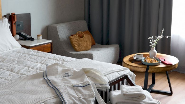 A cozy bedroom with a neatly made bed, robes, and towels. There's a chair, a small table with a vase, and a curtain by the window.