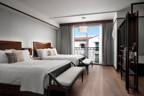 The image shows a hotel room with two neatly made beds, wooden furniture, and a window with a view of rooftops outside.