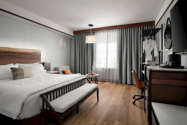 A modern hotel room with a bed, armchair, desk, TV, robe, and large window with curtains.