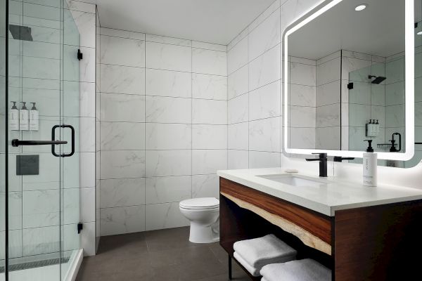 A modern bathroom featuring a glass shower, backlit mirror, wooden vanity, and white marble tiles.