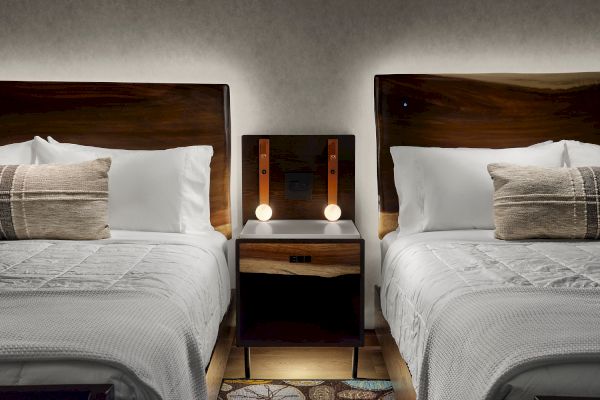 A cozy bedroom features two beds with white linens, brown pillows, and a central nightstand with two lamps and outlets.