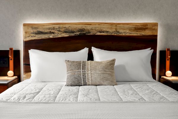 A neatly made bed with white pillows and a textured cushion is flanked by wooden nightstands and lamps, under a rustic wood headboard.