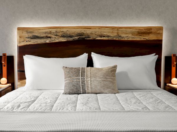 A neatly made bed with white pillows and a textured cushion is flanked by wooden nightstands and lamps, under a rustic wood headboard.