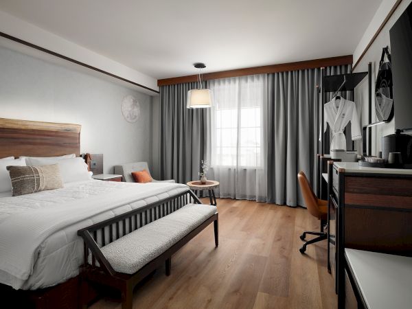 A modern hotel room with a large bed, bench, chair, desk, and TV. Gray curtains cover the window; a robe hangs next to a coffee setup.