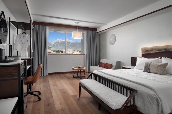 The image shows a modern hotel room with a large bed, bench, desk, sofa, and a view of mountains through the window.