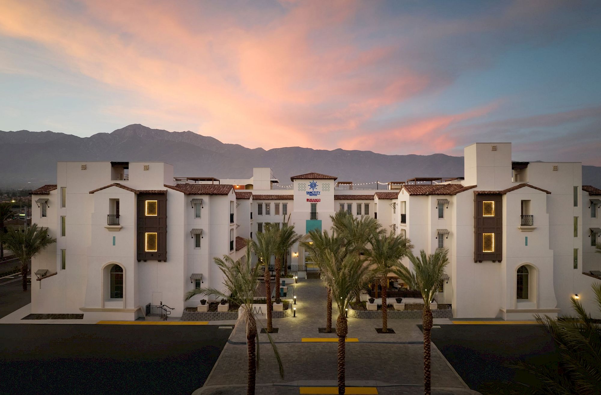 The image shows a white building with arches and palm trees in front, set against mountains at sunset.