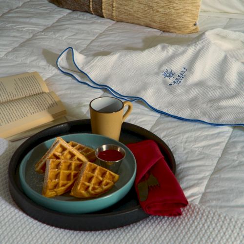 A cozy bed setup with a tray holding waffles, syrup, tea, a mug, an open book, and a pair of headphones on a white quilt.