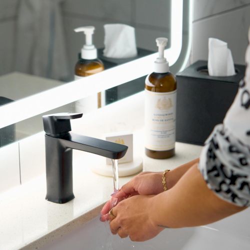 A modern bathroom sink with a black faucet, hands washing with water, and toiletries like soap dispensers and tissues on the counter, in a bright setting.