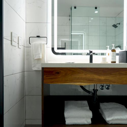 A modern bathroom with a wooden vanity, open shelf with towels, large mirror with backlit edge, towel on a rack, and a glass shower in the background.