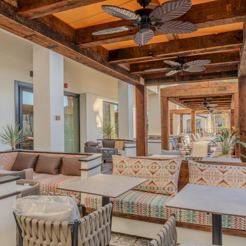 Outdoor seating area with patterned cushions, wooden beams, ceiling fans, and palm trees in a bright and airy setting.