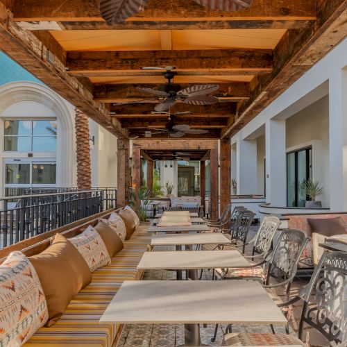 Outdoor dining area with empty tables and chairs, decorated with cushions. Overhead fans and wooden beams under a covered patio.