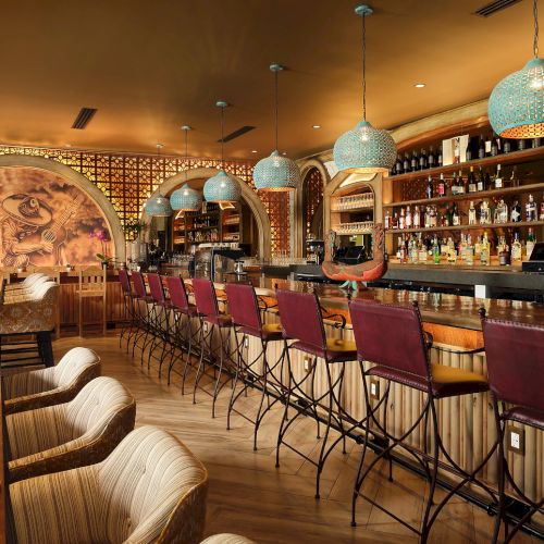The image shows a stylish bar with red chairs, ornate lighting, a mural, and a variety of bottles displayed on shelves.