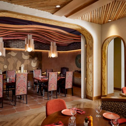 Dining area with wooden decor, patterned chairs, hanging lights, and intricate wall art; set tables with red napkins and glassware.