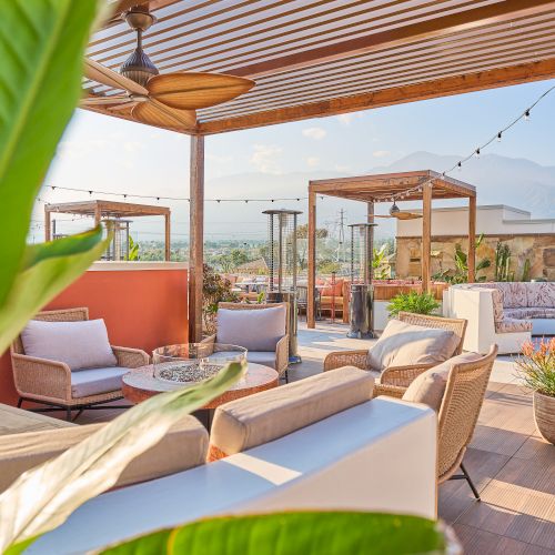 A rooftop patio with cozy seating, a pergola, green plants, and string lights creates a relaxing, outdoor lounge space.