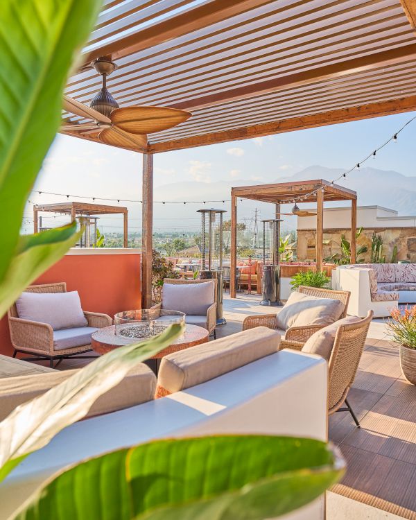A rooftop patio with cozy seating, a pergola, green plants, and string lights creates a relaxing, outdoor lounge space.