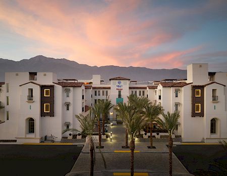 A large building with white walls and a central entrance is surrounded by palm trees, set against a mountain backdrop and colorful sky.