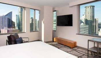 A modern hotel room with large windows offering a city view, a bed, chair, TV on the wall, and a bench under the TV, ending the sentence.