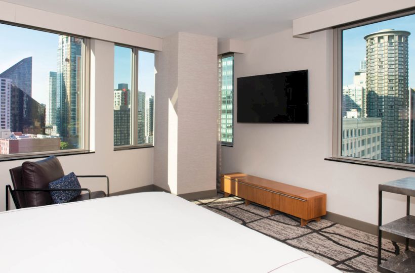 A modern hotel room with large windows offering a city view, a bed, chair, TV on the wall, and a bench under the TV, ending the sentence.