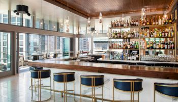 A modern bar with a wooden counter, blue stools, shelves stocked with bottles, and large windows offering city views, creating an inviting atmosphere.
