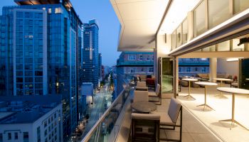 A modern cityscape at dusk with high-rise buildings, a lit-up street, and a balcony area featuring tables and chairs overlooking the urban view.