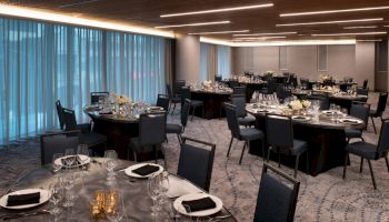A modern banquet hall with round tables set for a formal event, featuring elegant table settings, black napkins, and soft lighting from overhead fixtures.