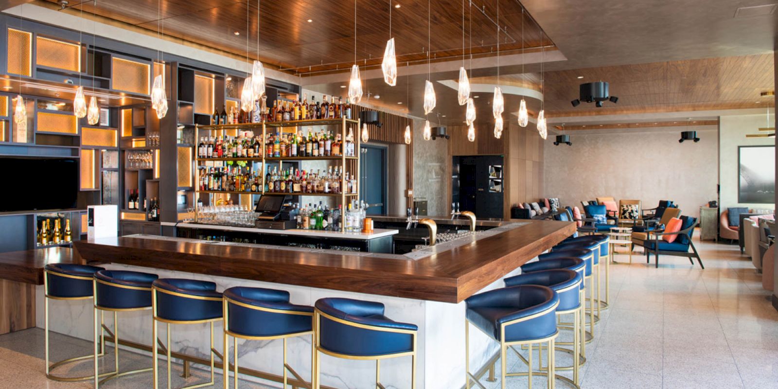 A modern bar with blue stools, a well-stocked shelf of beverages, hanging lights, a wooden ceiling, and a cozy seating area in the background.
