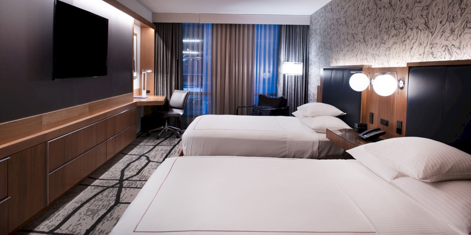 The image shows a modern hotel room with two beds, a desk, a flat-screen TV, and wall lights. The decor is minimalist with neutral tones.