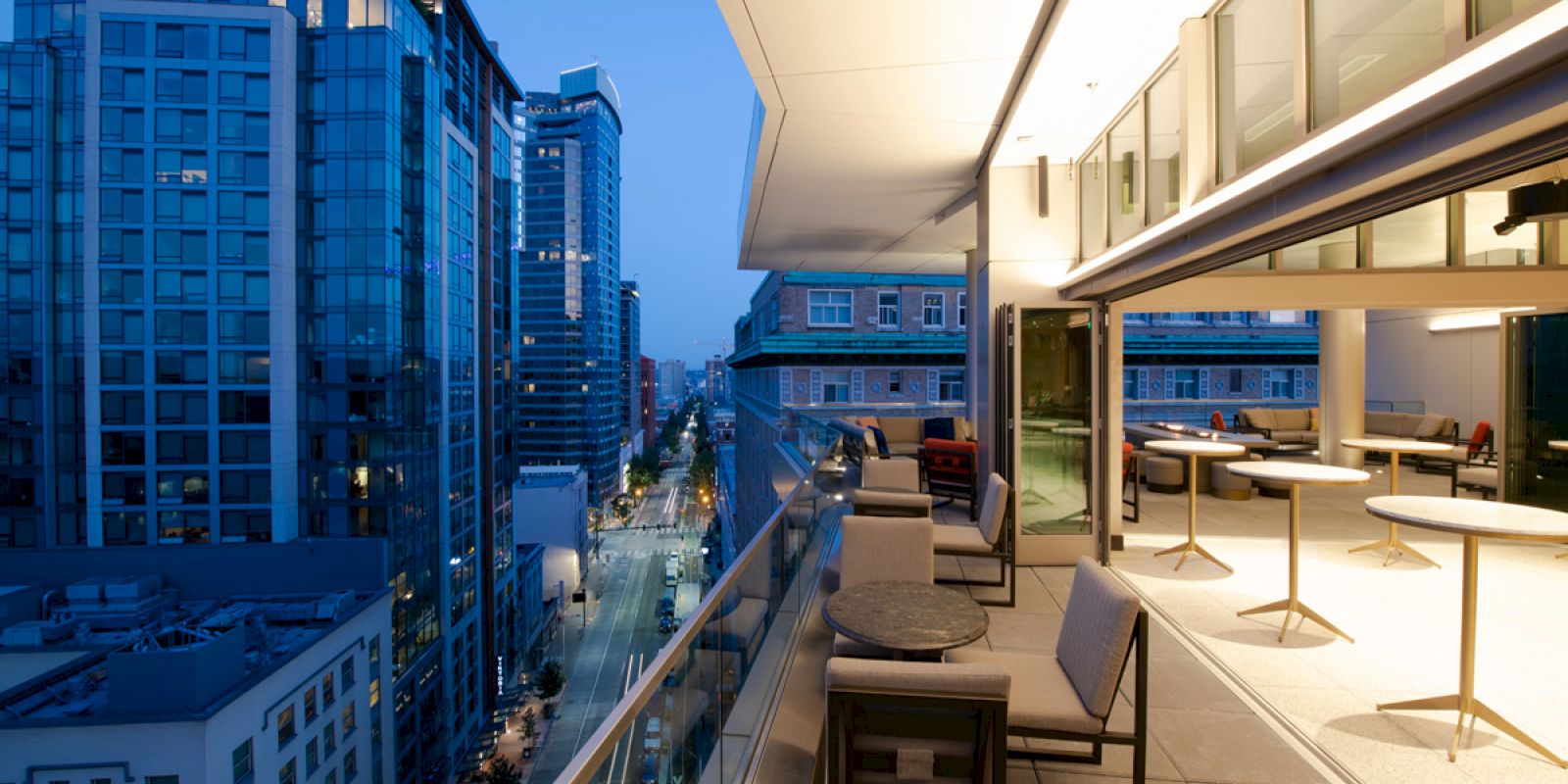 This image shows a modern balcony with seating area and tables, overlooking a city street and tall buildings at dusk.
