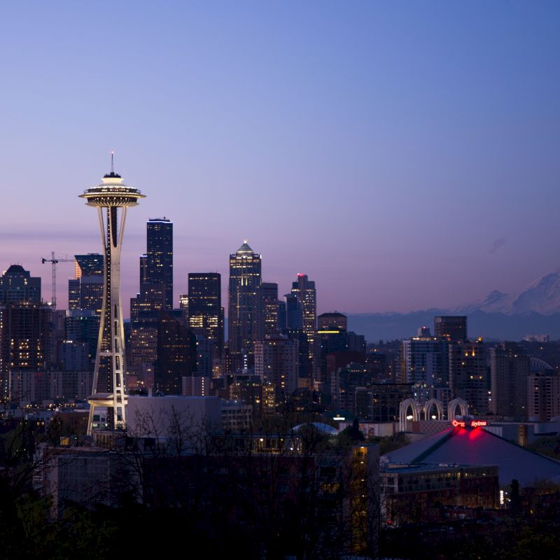 The image shows a nighttime cityscape of Seattle, featuring the Space Needle and a backdrop of downtown skyscrapers with Mount Rainier in the distance.