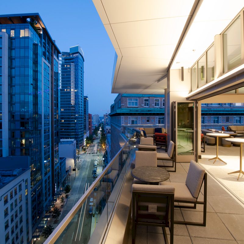 A modern cityscape at dusk, featuring high-rise buildings and a sleek balcony with seating and tables, overlooking a street below.