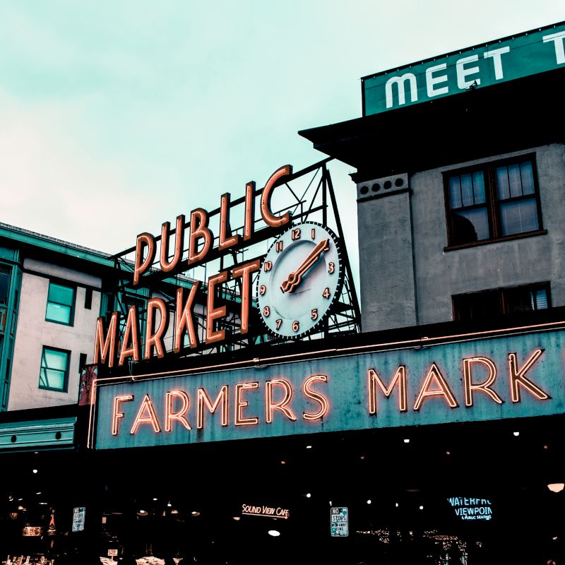 The image shows the iconic neon signs of a Public Market and Farmers Market against an urban backdrop with visible buildings and a cloudy sky.