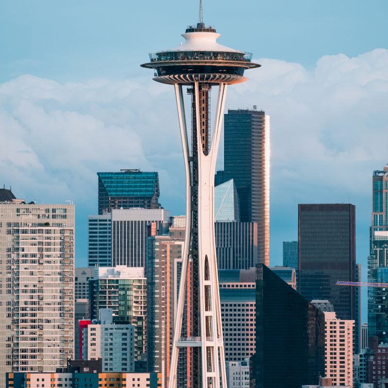 The image features the Space Needle surrounded by the Seattle skyline, showcasing a mix of modern high-rise buildings against a backdrop of clouds.