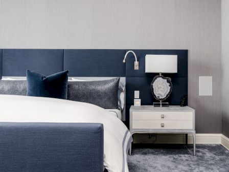A modern bedroom with a navy blue headboard, white bed linen, a bedside table with a decorative lamp, and a gray carpet floor.